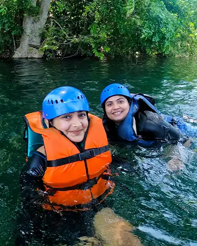 Two happy women floating in the fresh, green river water during a swimming or river crossing activity in Dandeli, wearing safety gear.